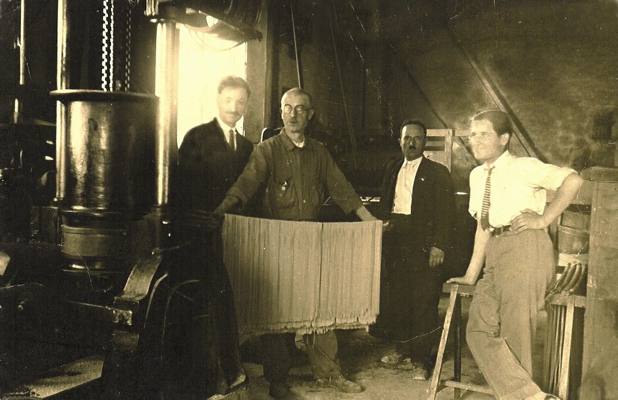 Four men in a pasta workshop beside a large press. The man in the middle holds up a curtain of fresh long pasta.
