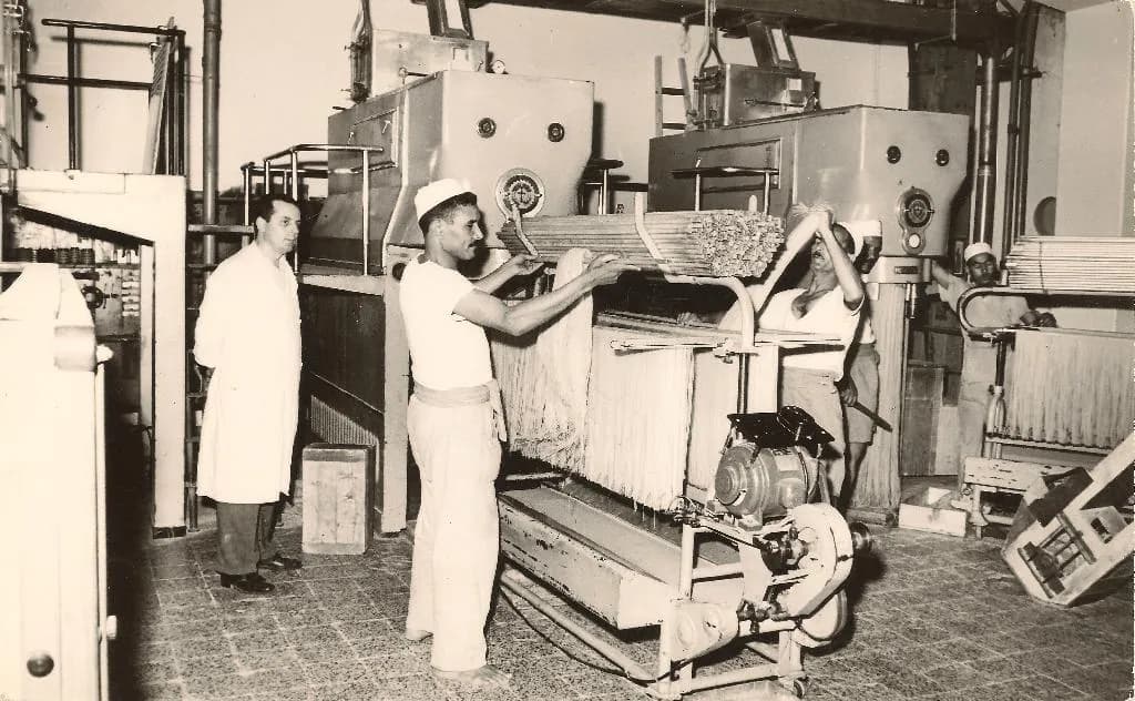 Workers in white coats at pasta machines on a factory floor.