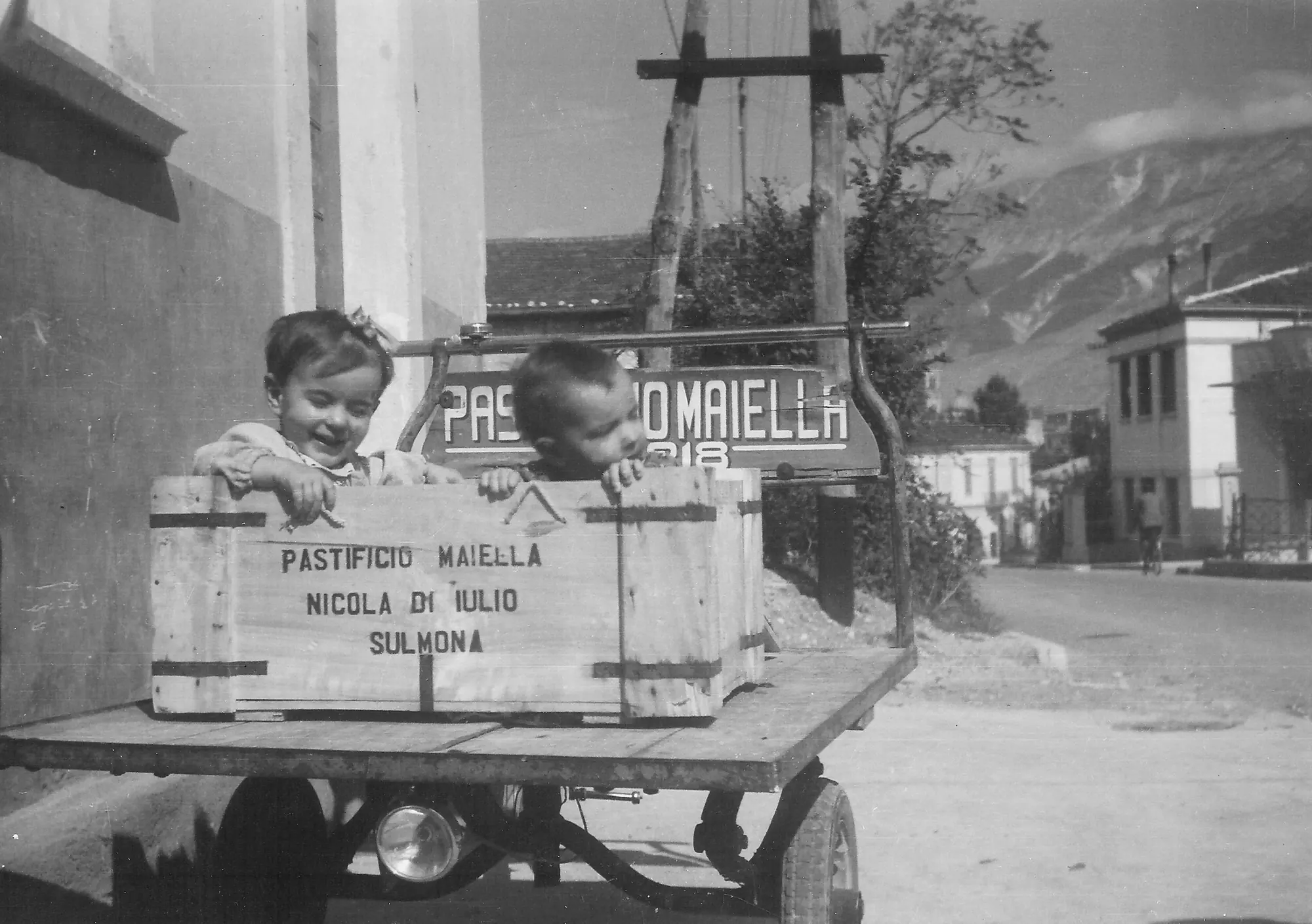 Two small children sit in a wooden crate stenciled Pastificio Maiella, Nicola Di Iulio, Sulmona, on a delivery cart in an Italian street.