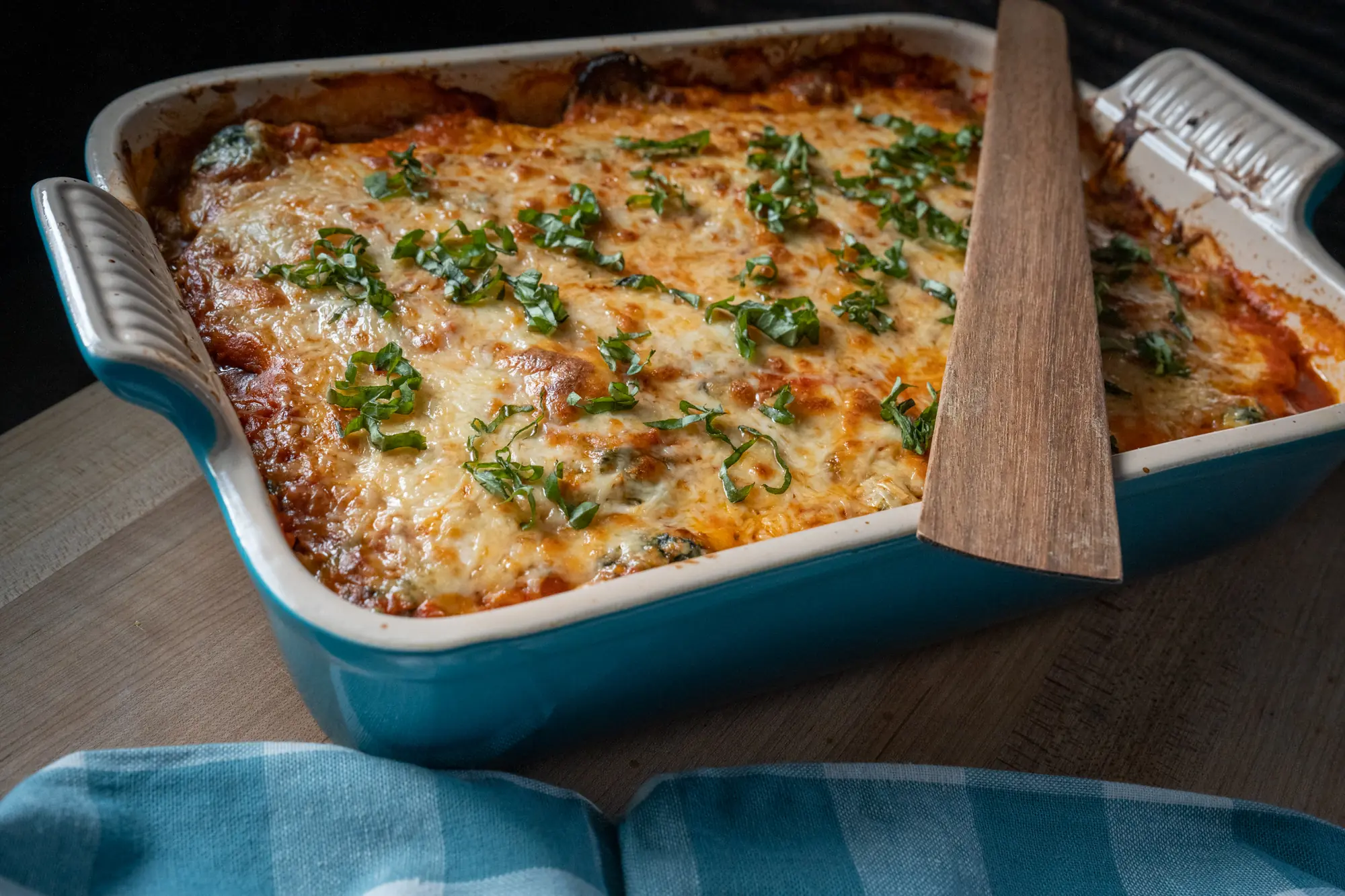 Polenta lasagna in a blue baking dish, browned and bubbly, topped with fresh basil, a wooden spatula resting in the pan.
