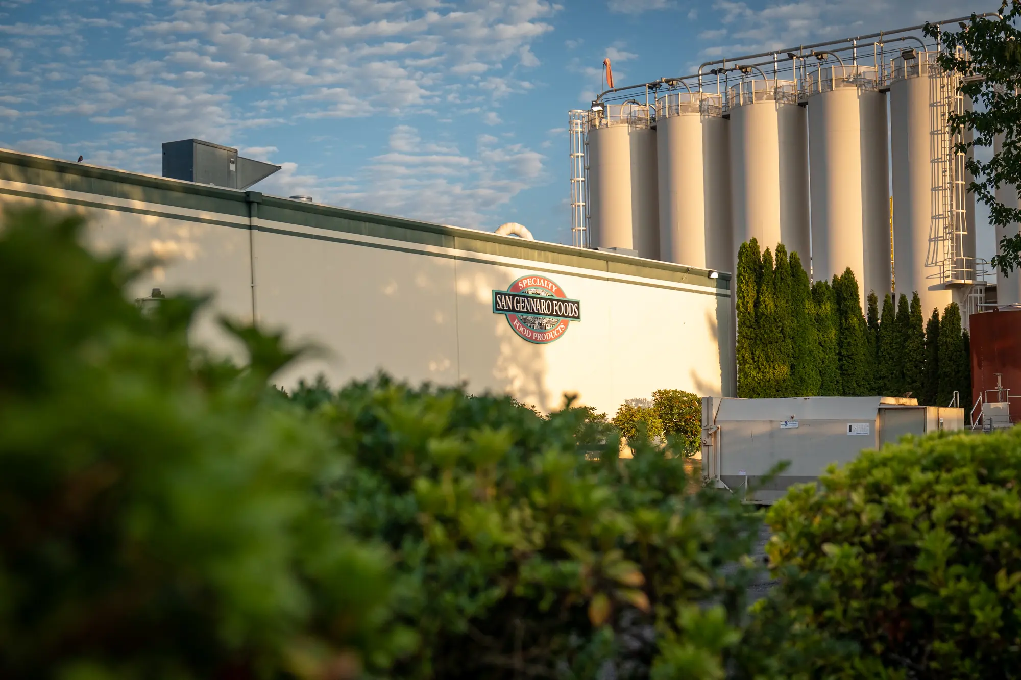 The San Gennaro Foods building, its logo on the wall and grain silos behind it, seen past green shrubs.
