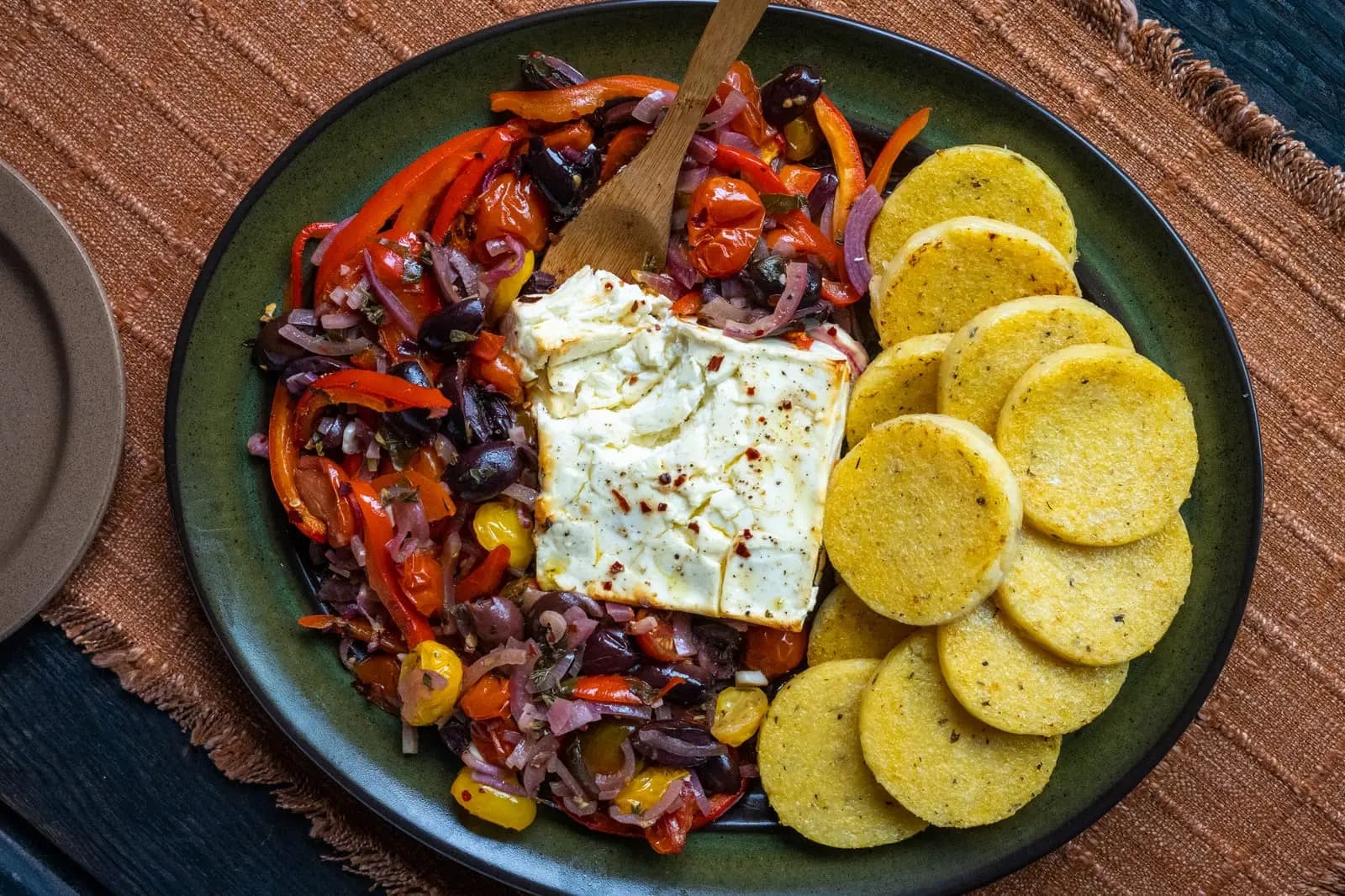 Baked feta with roasted peppers, cherry tomatoes, olives and red onion beside golden polenta rounds on a green platter.