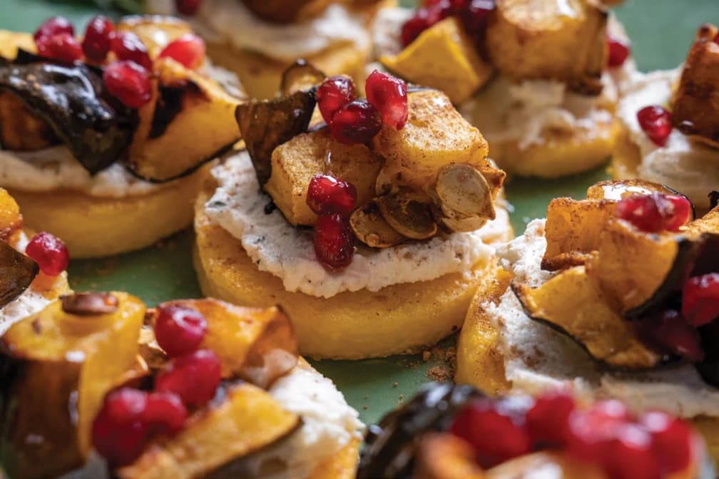 Polenta rounds topped with whipped cheese, roasted squash, squash seeds and pomegranate on a green plate.