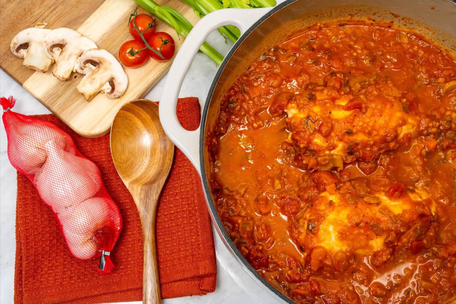 Chicken simmering in cacciatore stew in a white pot, beside mushrooms, tomatoes and celery.