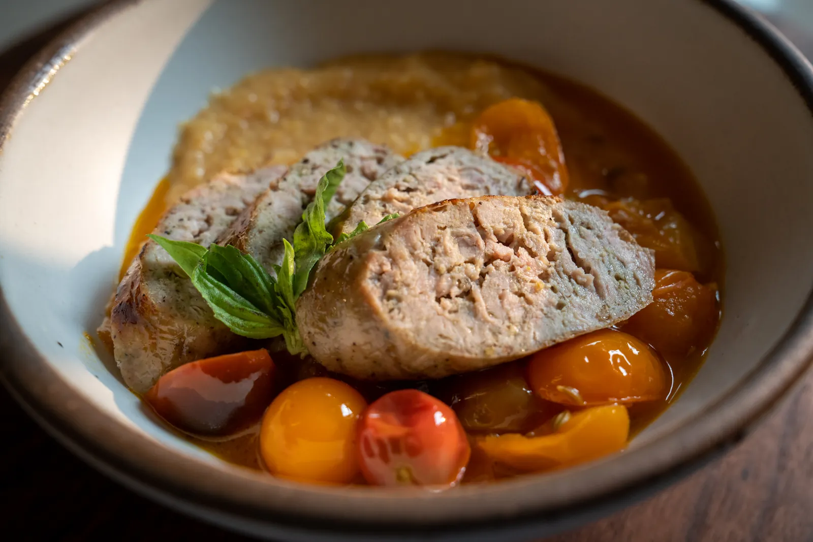 Creamy polenta with sliced grilled sausage, blistered cherry tomatoes and basil in a white bowl.