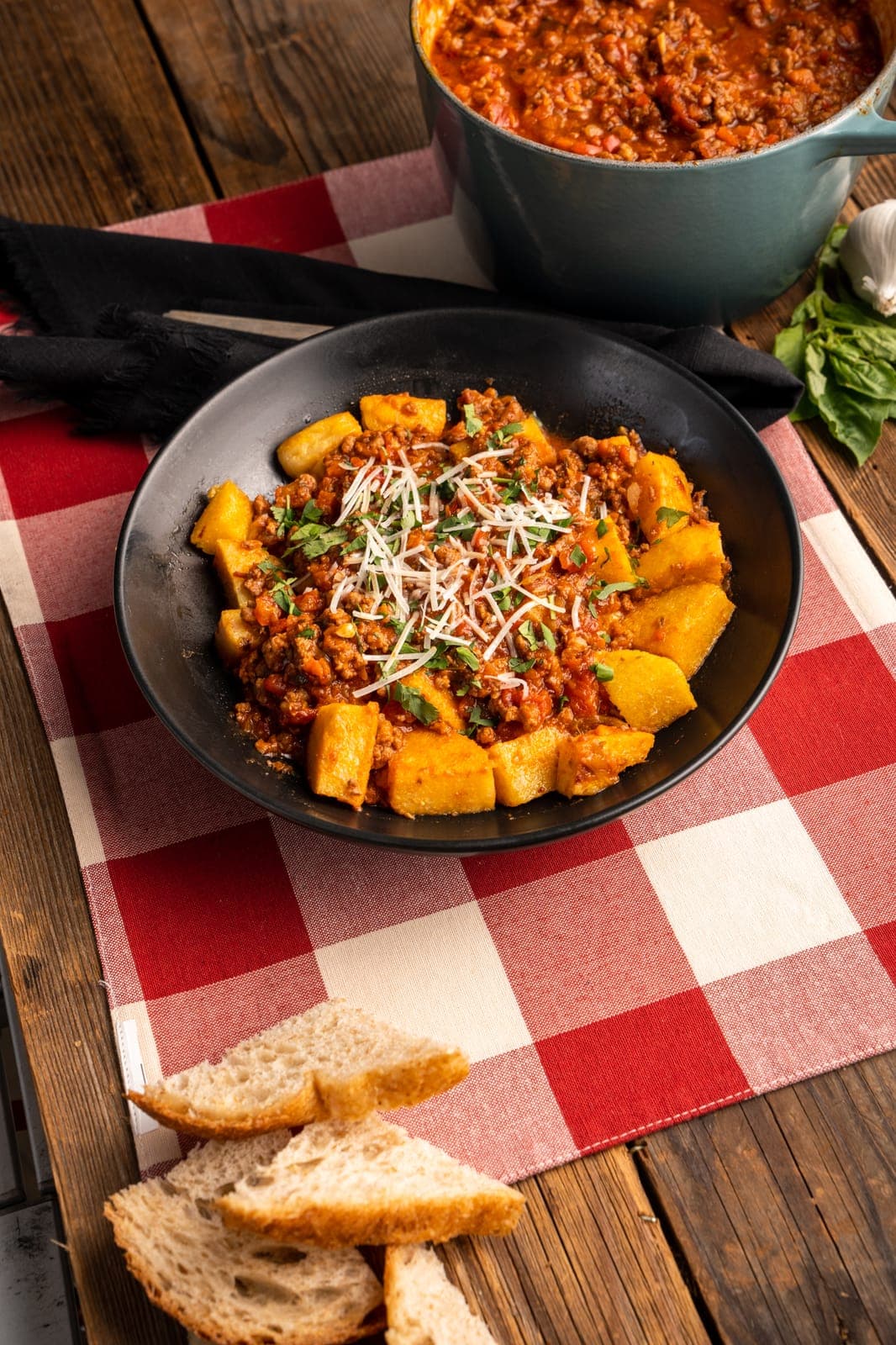 Golden polenta cubes and meat sauce topped with shredded parmesan and parsley in a black bowl, with a pot of sauce and sliced bread.