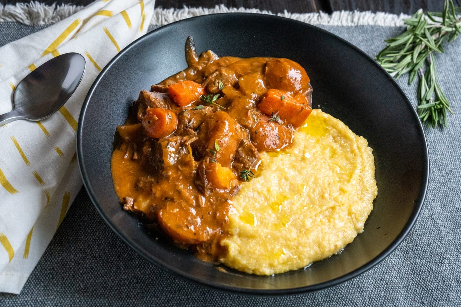Beef stew with carrots and thyme beside creamy polenta drizzled with olive oil in a black bowl, a sprig of rosemary nearby.