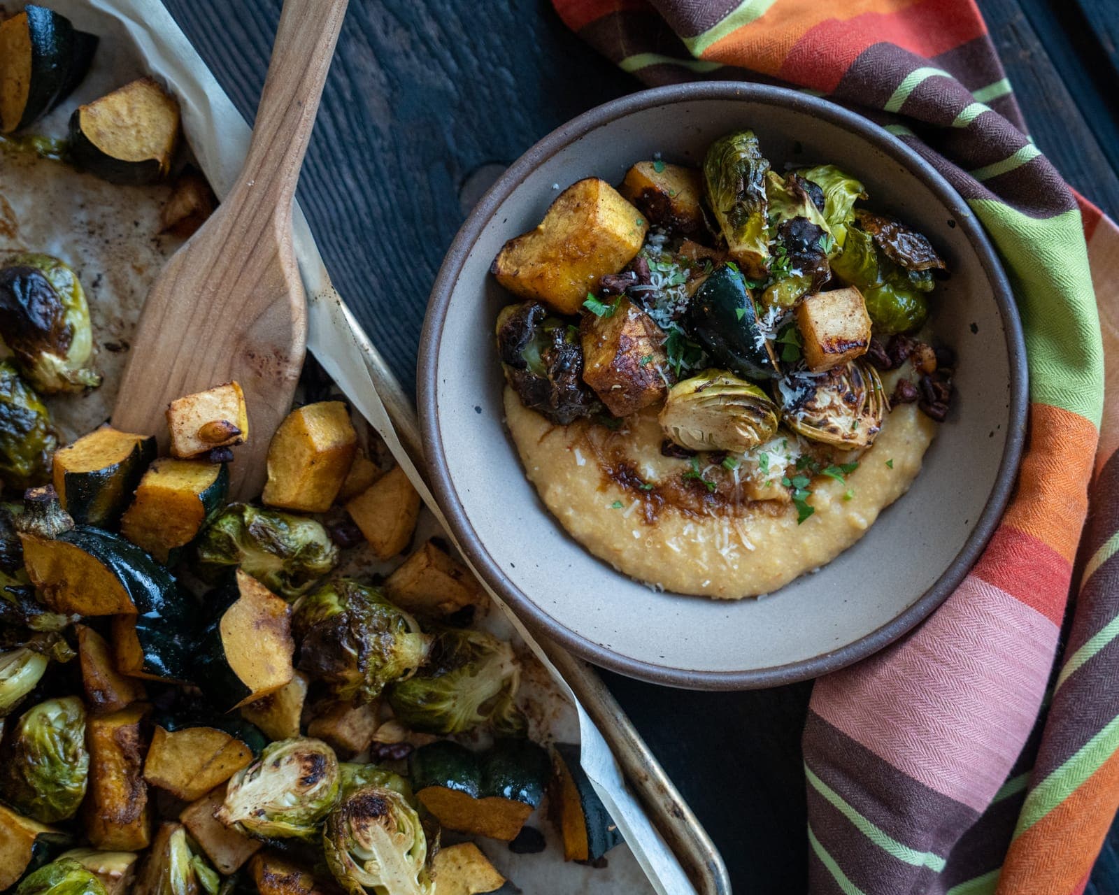 Creamy polenta topped with roasted squash, Brussels sprouts and grated parmesan in a stoneware bowl, beside a sheet pan of roasted vegetables.